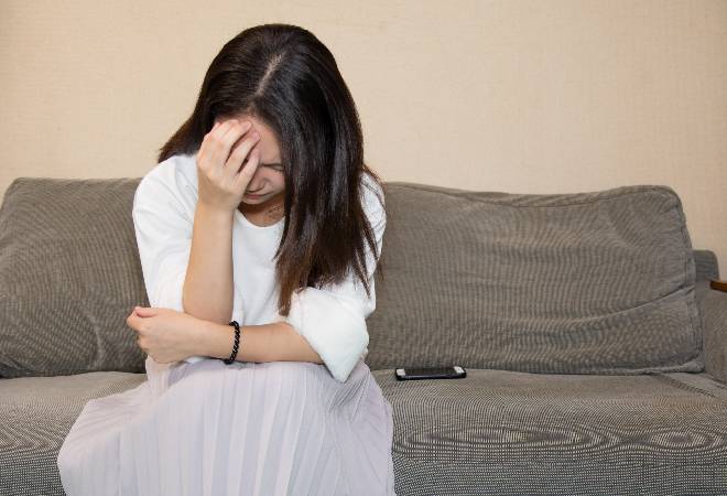 スマホをソファーの上に置き、項垂れる女性の姿。